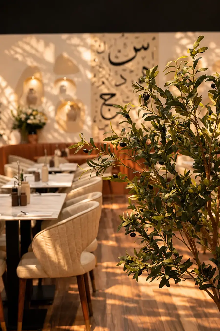An olive tree beside cream dining chairs and tables, the calligraphy wall and lit niches behind.