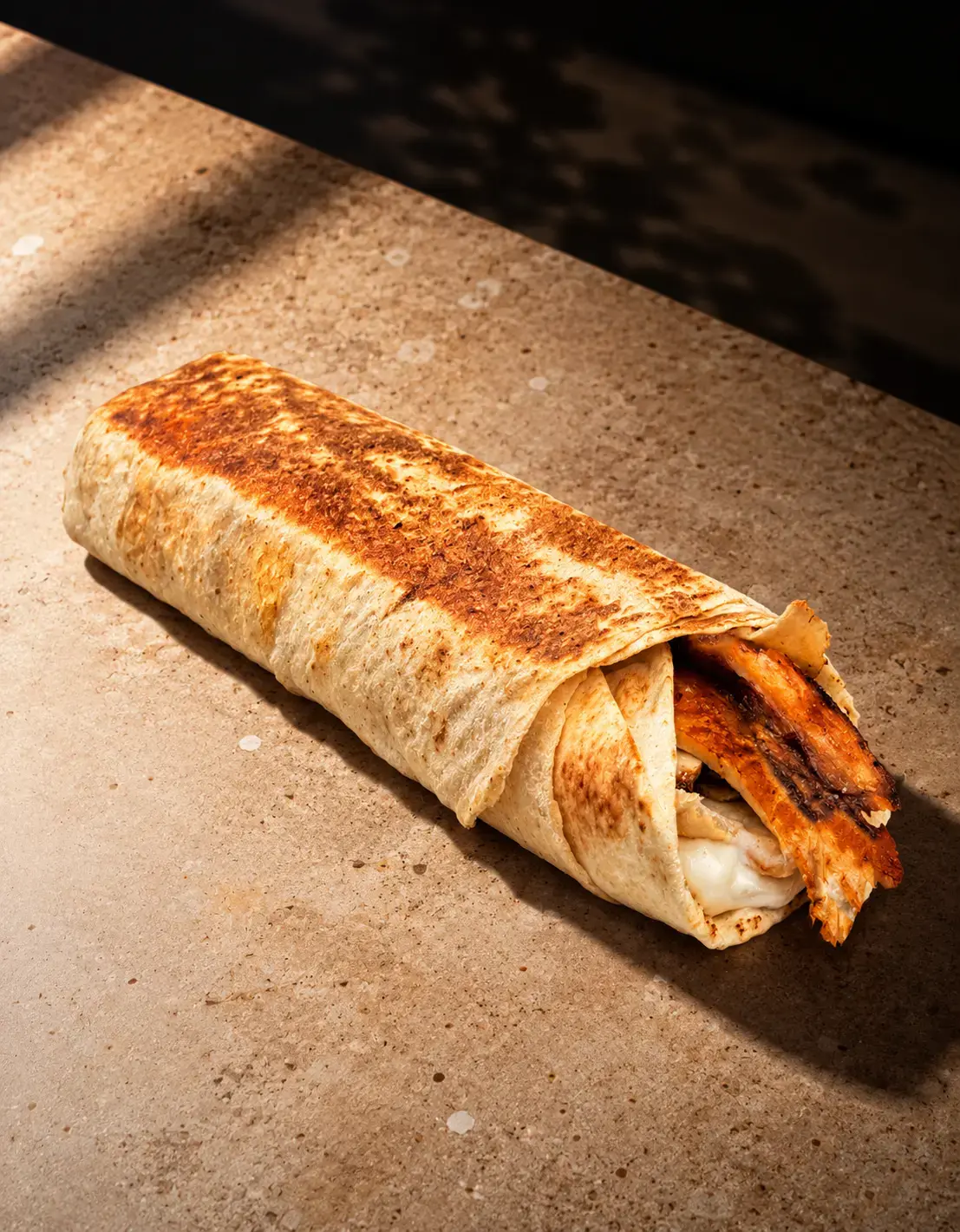 A chicken shawarma saj wrap, toasted golden, with garlic sauce and charred chicken at the open end, on a stone counter.