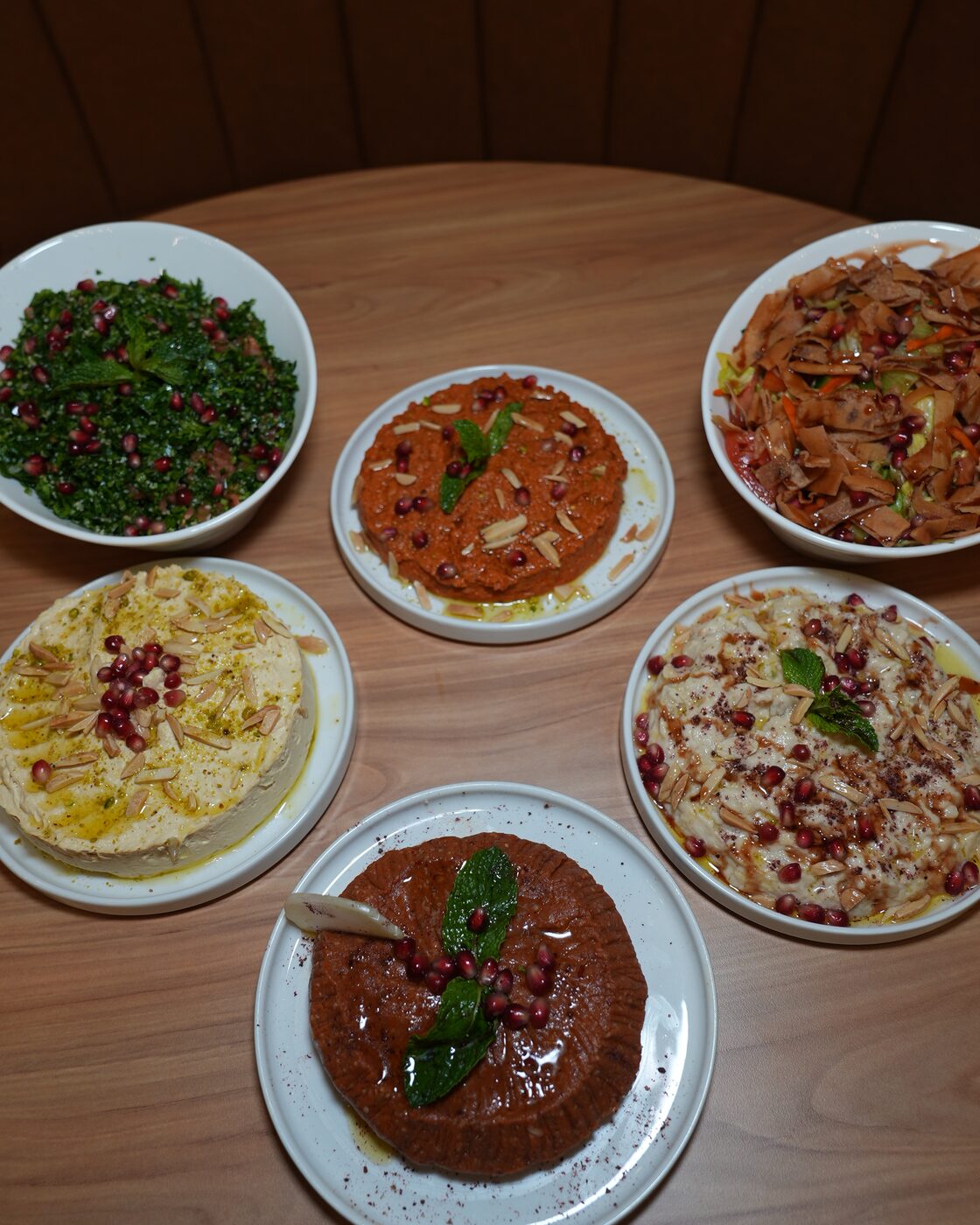 A round table set with a spread of mezza: tabbouleh, muhammara, fattoush, hummus, moutabal and a sumac-dusted dip, each finished with pomegranate seeds, almonds and mint.